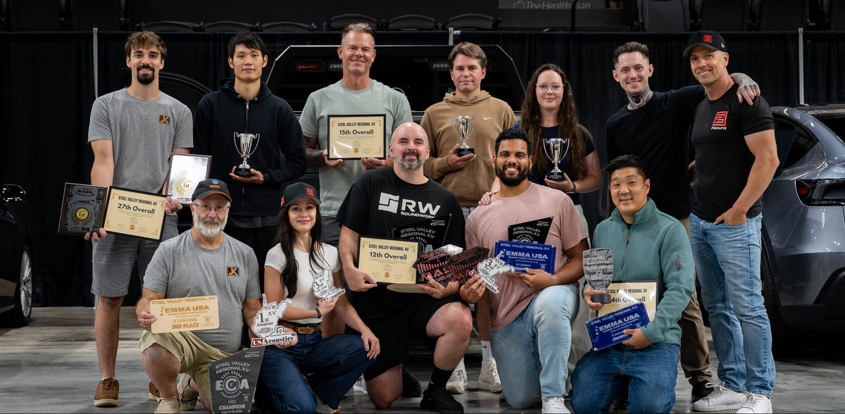 Group of people holding certificates and trophies in an automotive workshop with vehicles in the background