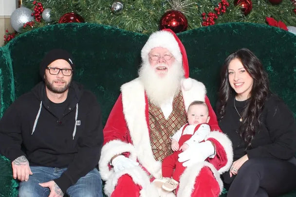 Santa Claus seated with a baby and two adults on a green couch, surrounded by festive Christmas decorations and greenery