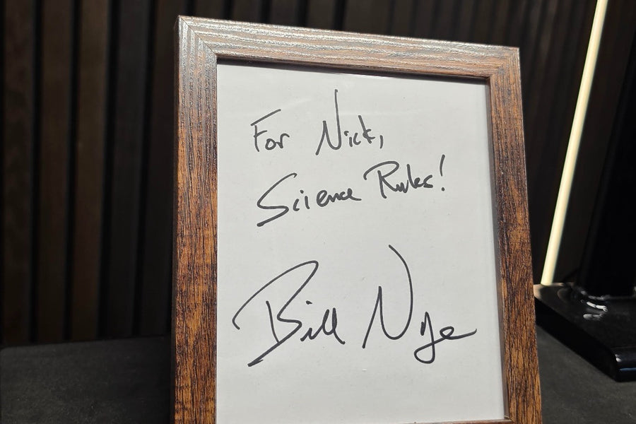 Framed handwritten note reading For Nick Silence Rules and signed Bill Nye, displayed on a dark wooden surface beside a light colored cable