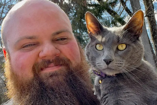 Bearded man posing outdoors with a gray cat, both looking at the camera with trees in the background