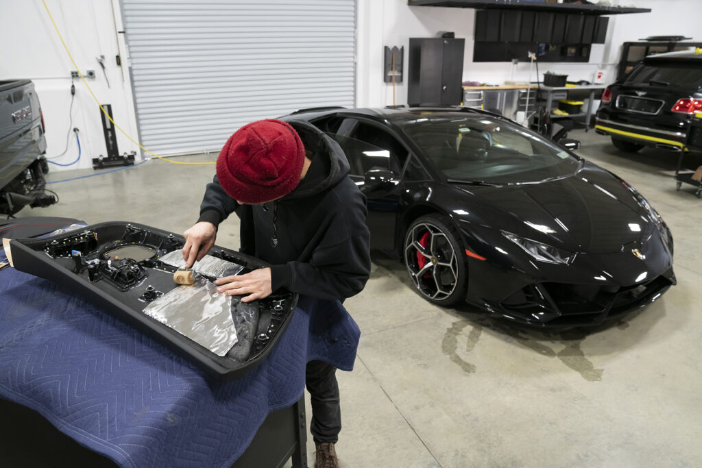 Person installing ResoNix CLD Squares sound deadening material onto the back of a lamborghini huracan's door panel to reduce rattles and distortion in a sound system installation with the lamborghini huracan in the background